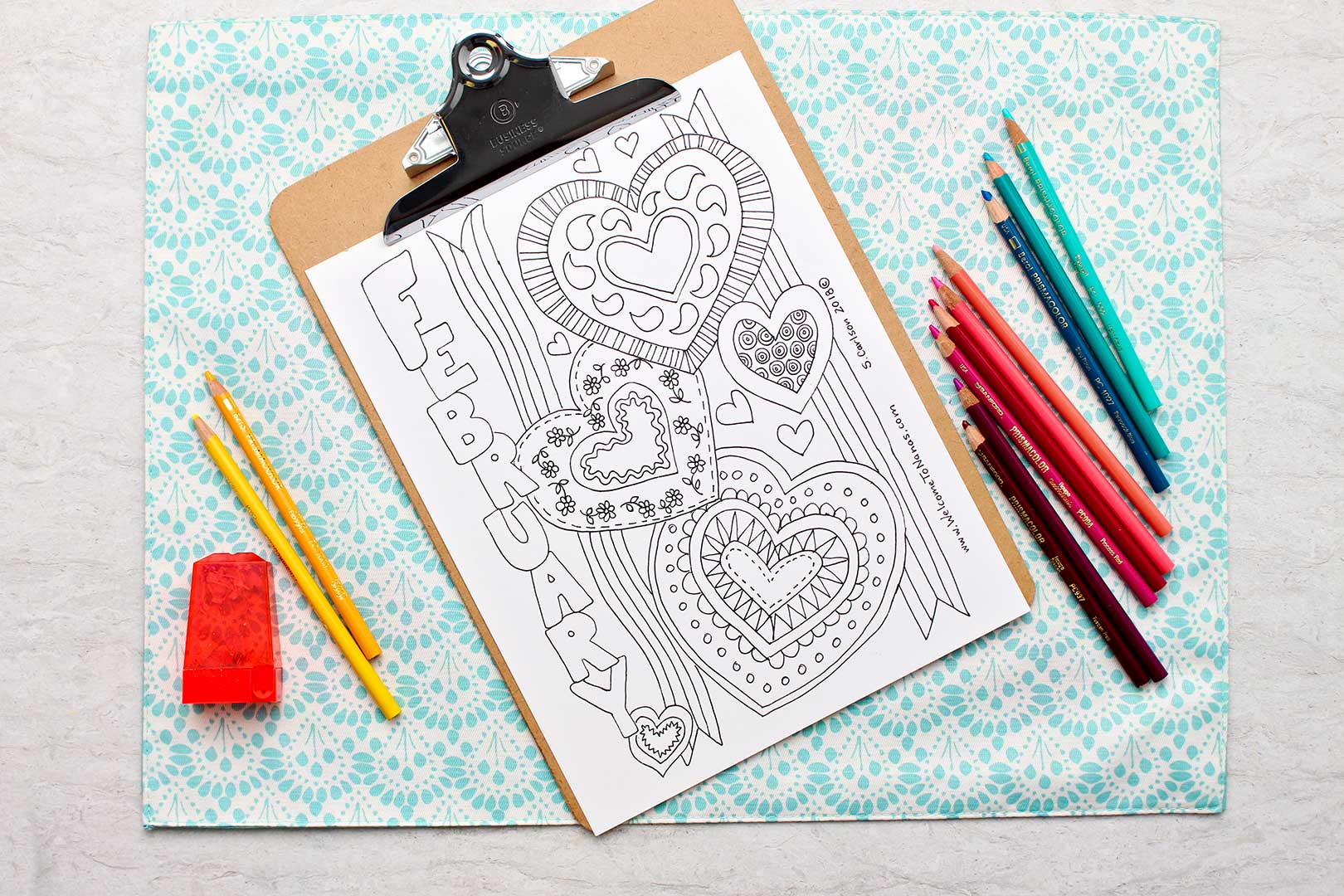 Uncolored February Coloring Page on clipboard with colored pencils near by.