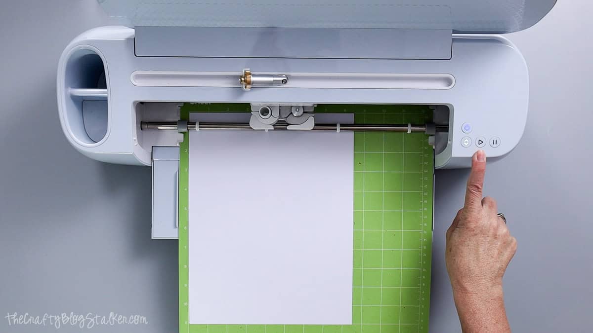 Cricut cutting machine cutting white cardstock on a green cutting mat while a hand presses the start button.