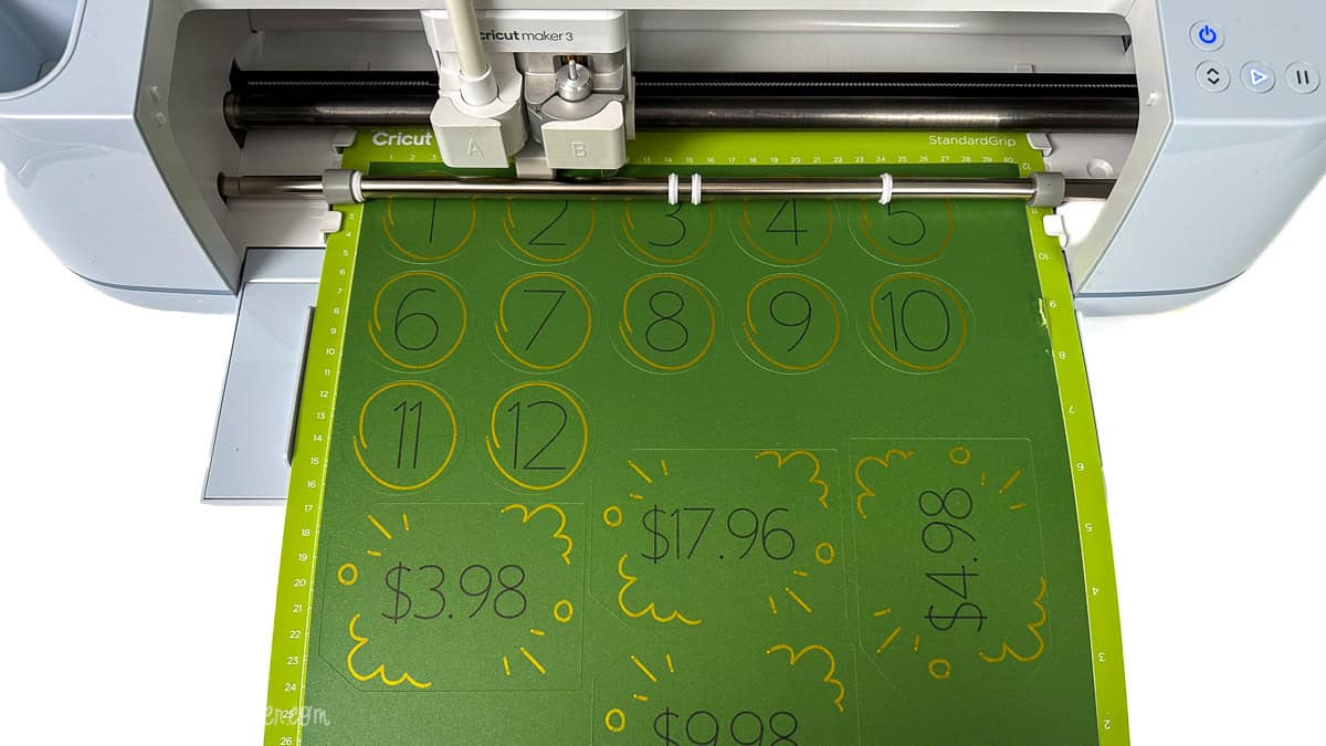 Cricut Maker 3 cutting machine drawing numbered circles and price tag designs on cardstock using a Cricut pen for a DIY Price Is Right baby shower game.