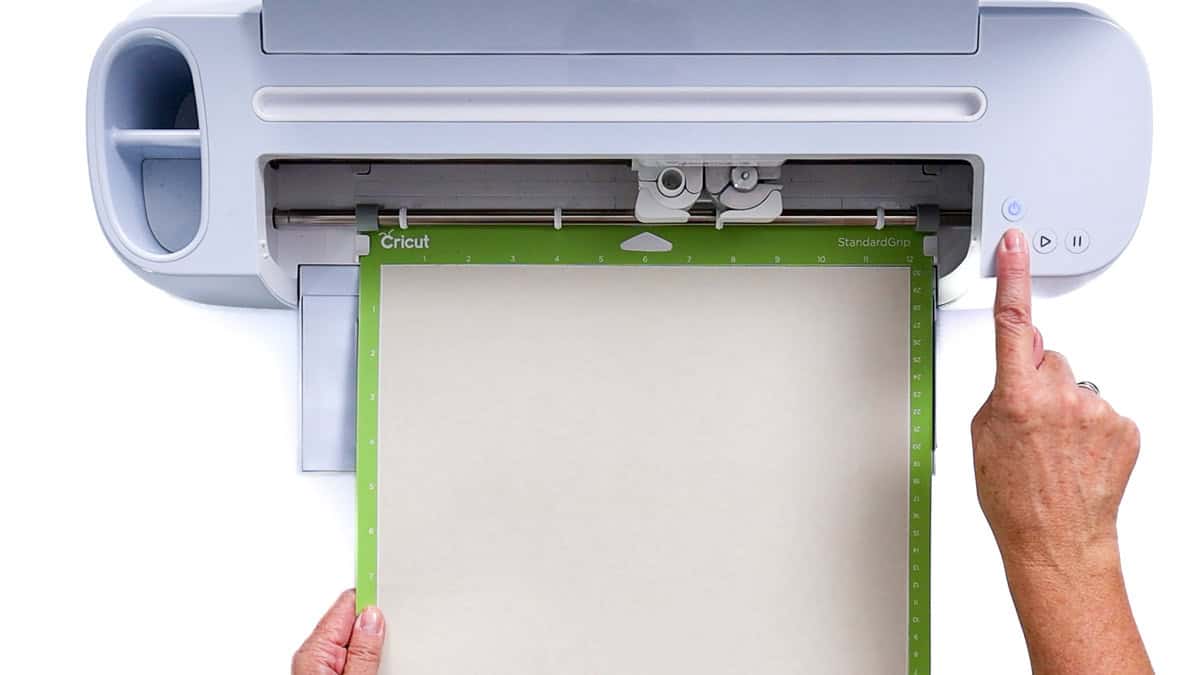 Hands load a sheet onto a Cricut cutting machine mat and press the start button to begin cutting a vinyl design.