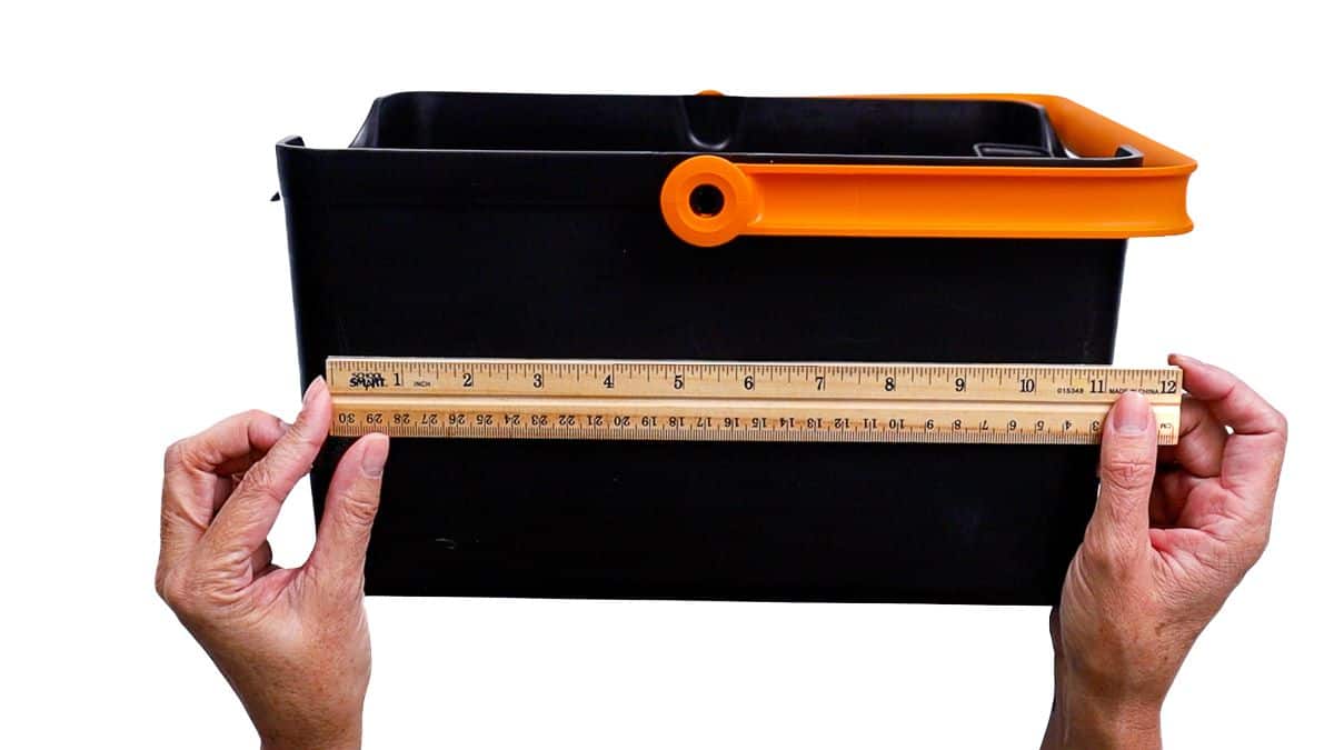 Hands holding a ruler across the front of a rectangular garden bucket to measure its width.