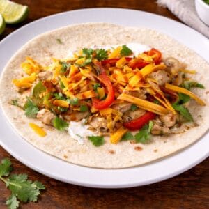Open flour tortilla filled with baked chicken fajitas, sautéed peppers and onions, shredded cheese, cilantro, and sour cream on a white plate.