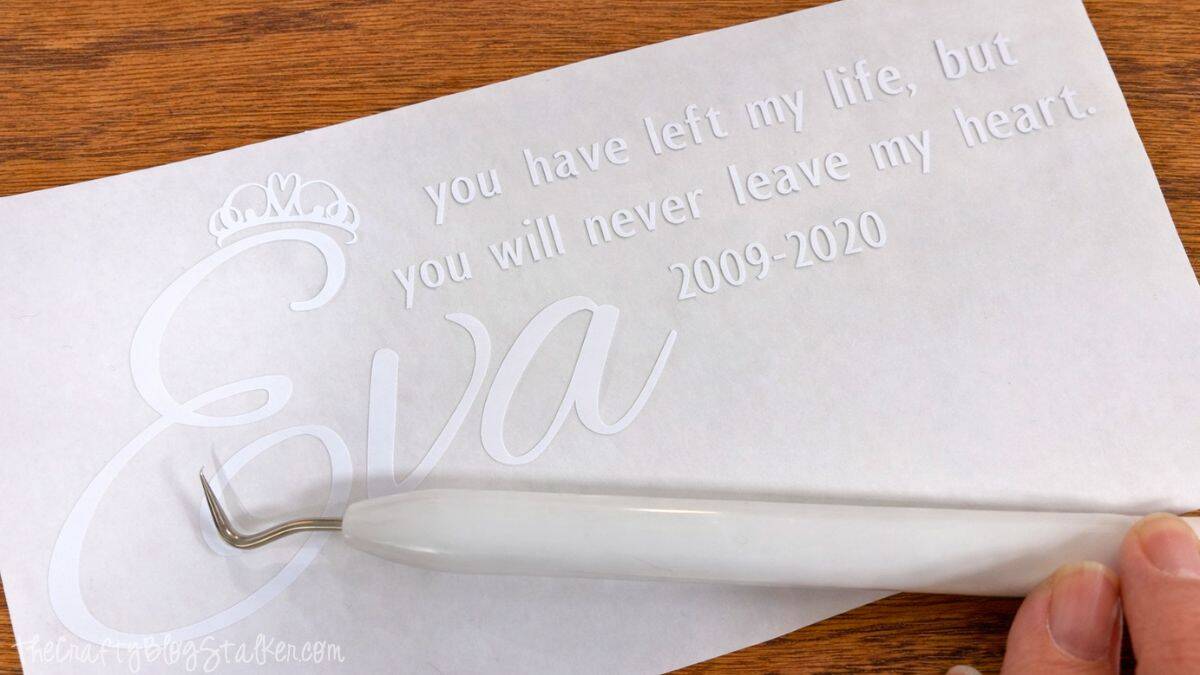 Weeded vinyl memorial design on transfer paper with a hook weeding tool lifting excess vinyl from a personalized pet remembrance quote and name.