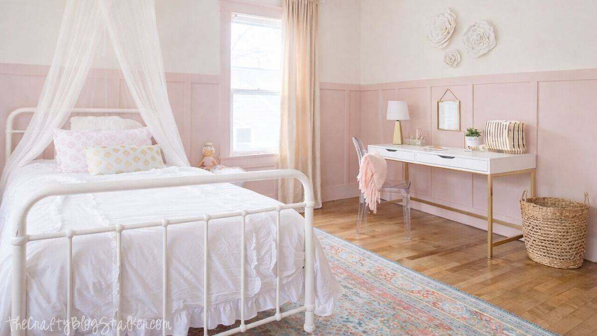 Styled girls bedroom with a white metal bed and canopy, window curtains, a desk with chair, wall decor, woven basket, and a patterned rug on hardwood floors.