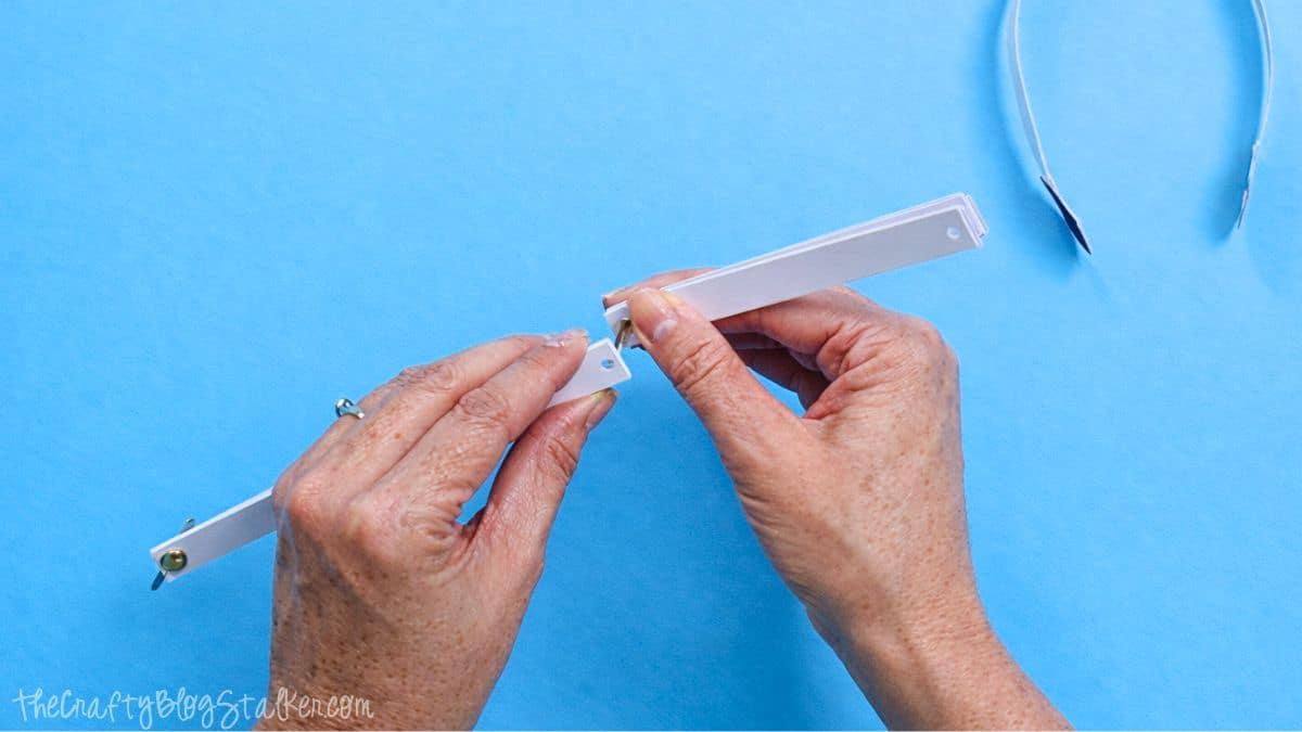 Hands connecting two white paper strips with a metal brad fastener, forming a joint for a paper strip snowman craft on a blue work surface.