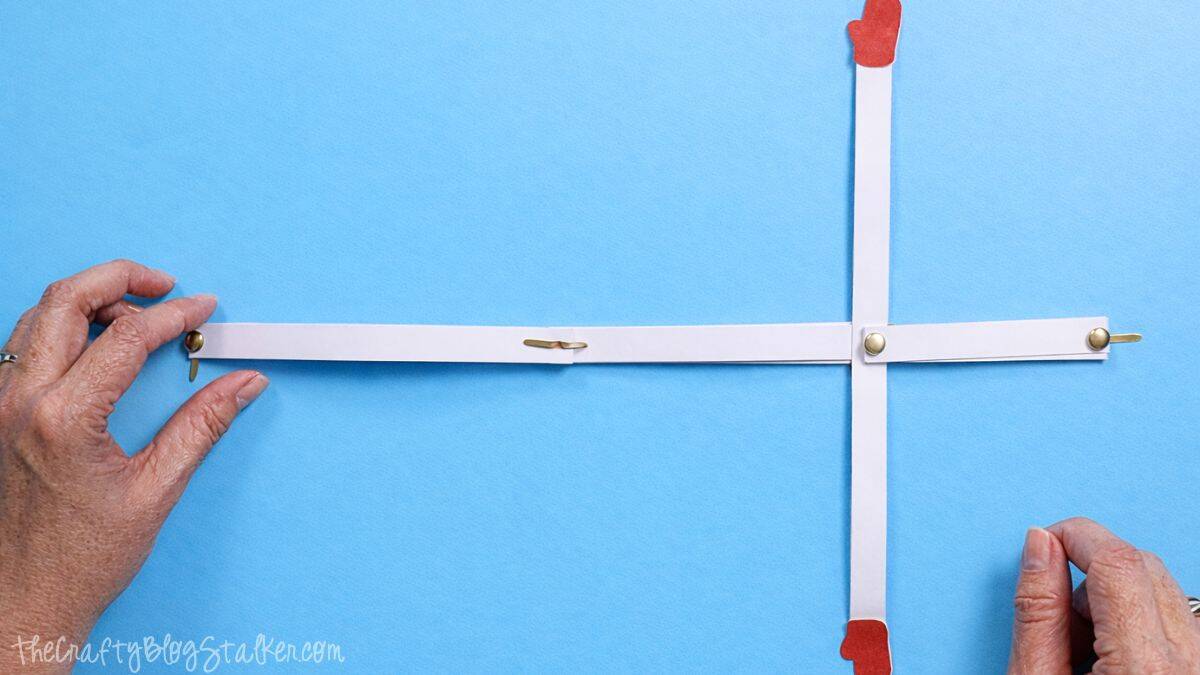 Hands arranging white paper strips joined with metal brads in a cross shape, with mitten cutouts on the ends, forming the snowman arm and body frame.