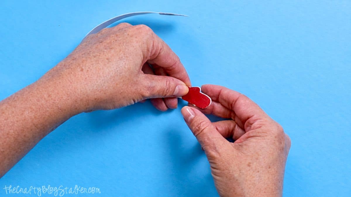 Hands attaching a small paper mitten to a curved paper strip, assembling parts of a paper strip snowman craft on a work surface.