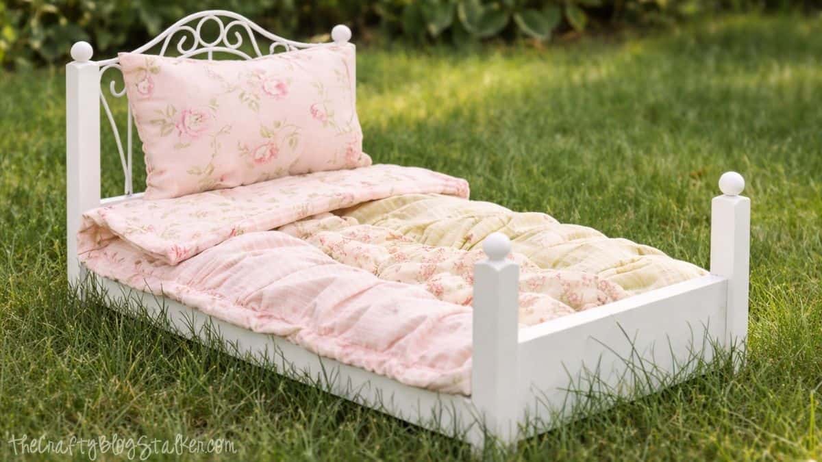 Handmade ruffled quilt and pillow on a small white doll bed placed on grass, showing a finished doll bedding set outdoors.