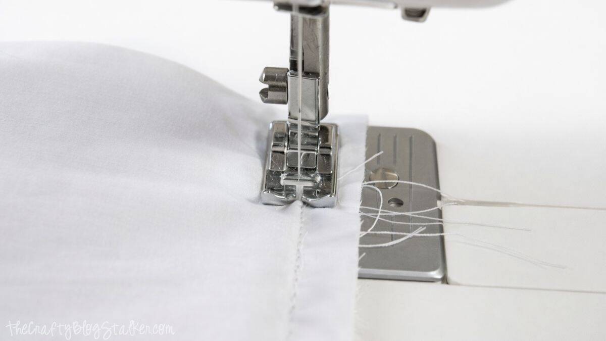 Close-up of a sewing machine foot stitching a straight seam along the edge of fabric, showing thread tails and the needle in action.