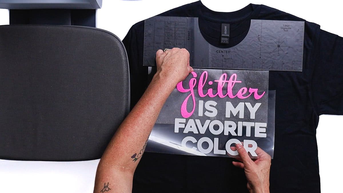 Hands centering an iron-on vinyl design on a T-shirt using a T-shirt alignment ruler, with a heat press platen positioned beside the shirt.