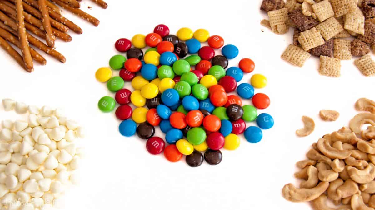 Ingredients for white chocolate party mix arranged on a white surface, including pretzels, cereal squares, white chocolate chips, cashews, and candy-coated chocolates.