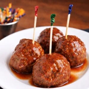 Sweet chili meatball appetizers on a white plate, each topped with a toothpick and coated in a sticky sauce, with extra picks blurred in the background.