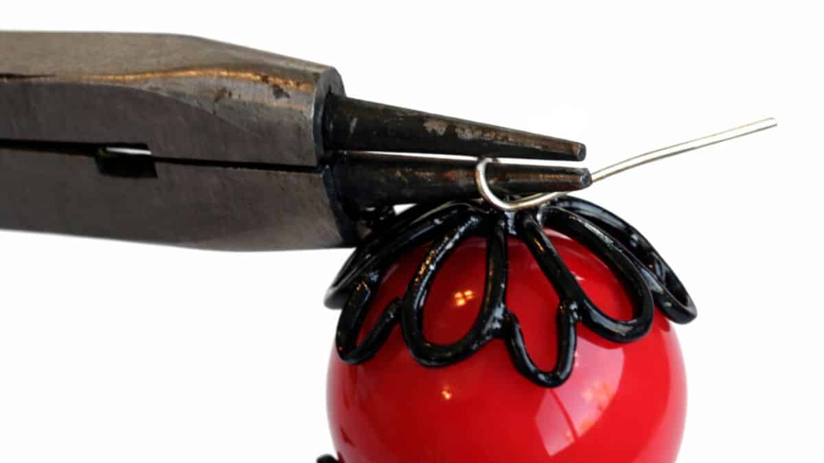 Pliers bending a wire loop at the top of a beaded spider body to form a hanger during assembly of a handmade spider craft.