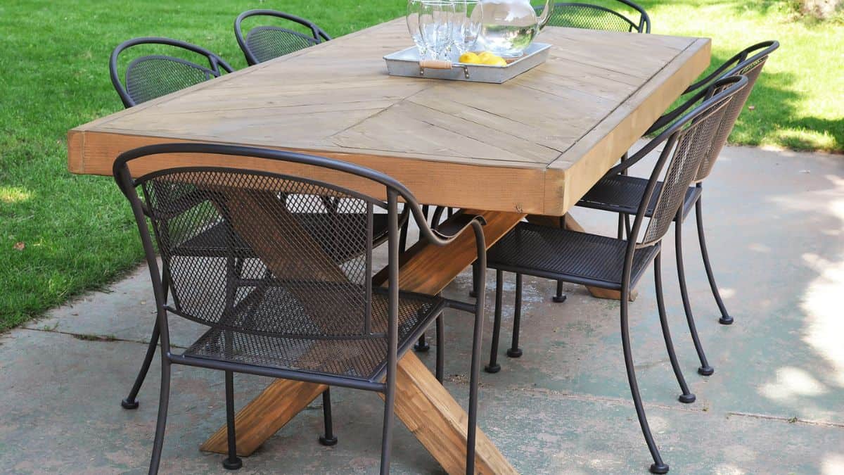 Handmade wooden outdoor dining table with a herringbone top and X-style base, surrounded by metal patio chairs on a concrete patio.