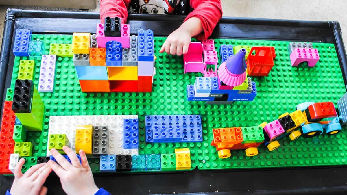Children playing with LEGO bricks on a DIY LEGO table, building colorful structures and vehicles on a large green baseplate.