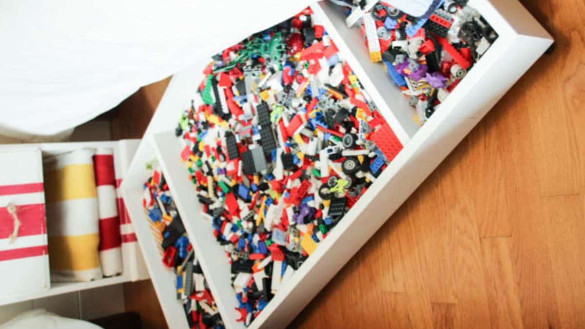 Under-bed LEGO storage drawer divided into sections, filled with sorted LEGO bricks, minifigures, baseplates, and boxed LEGO sets for organized play.