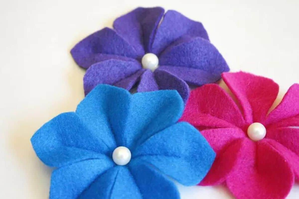 Three handmade felt camellia flowers with layered petals and pearl-style centers arranged on a light background as DIY craft embellishments.