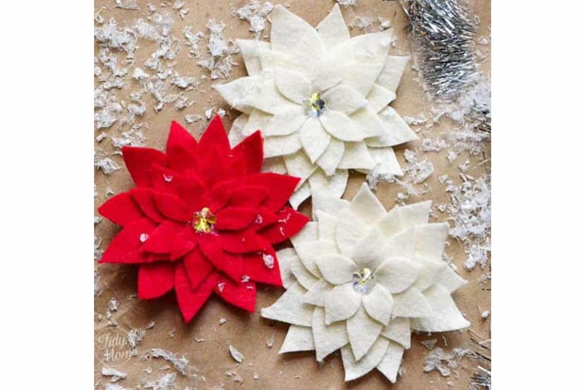 Handmade felt poinsettia flowers arranged on a rustic surface with faux snow, featuring layered petals and decorative centers for holiday decor or accessories.