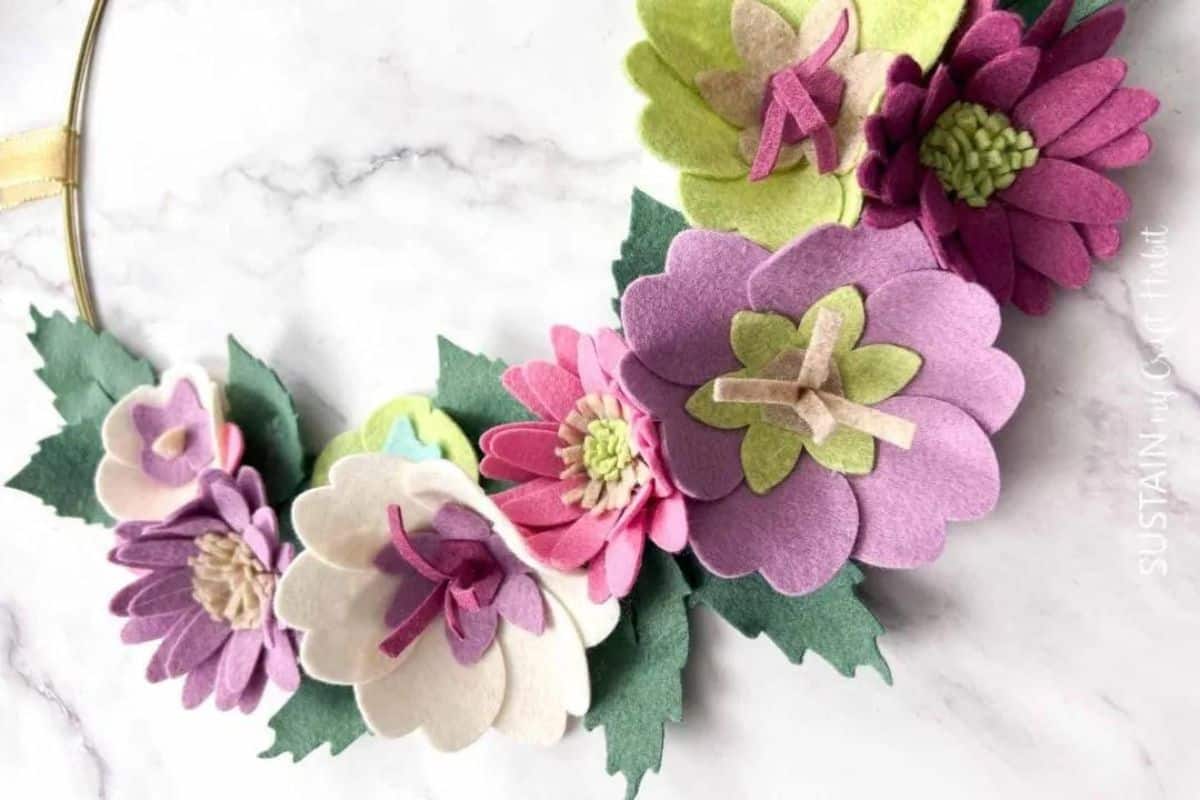 Gold hoop wreath decorated with handmade felt flowers and layered leaves, arranged in a floral cluster on a marble-style surface.