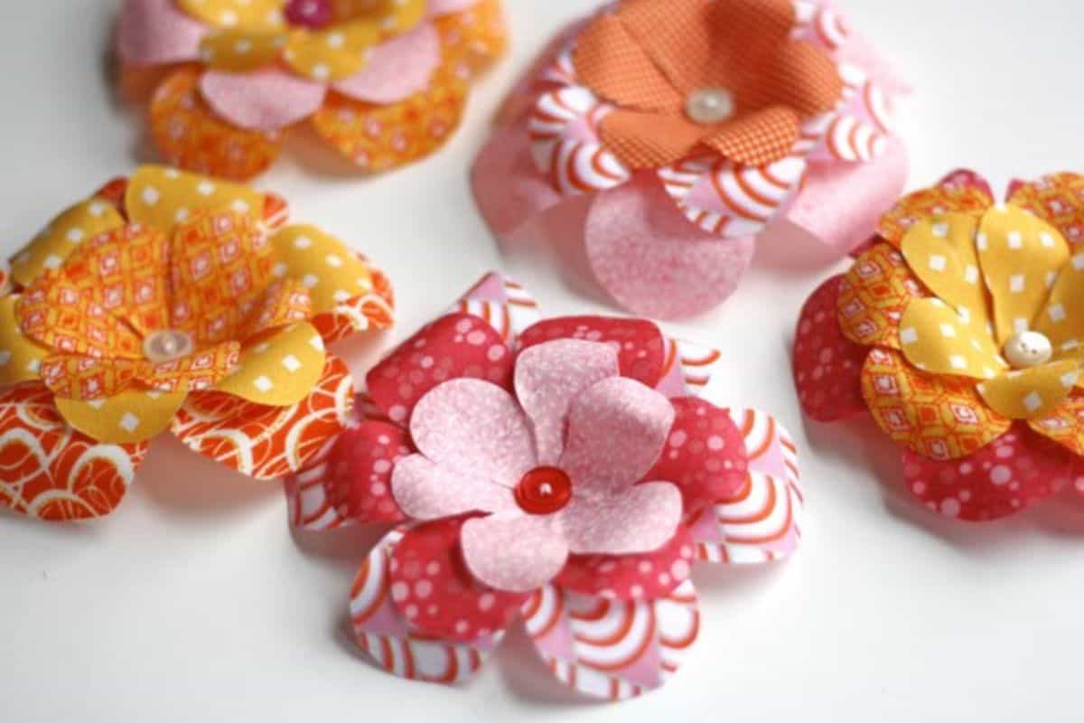A group of handmade fabric flowers with layered petals and button or bead centers, arranged together on a white surface as a craft display.