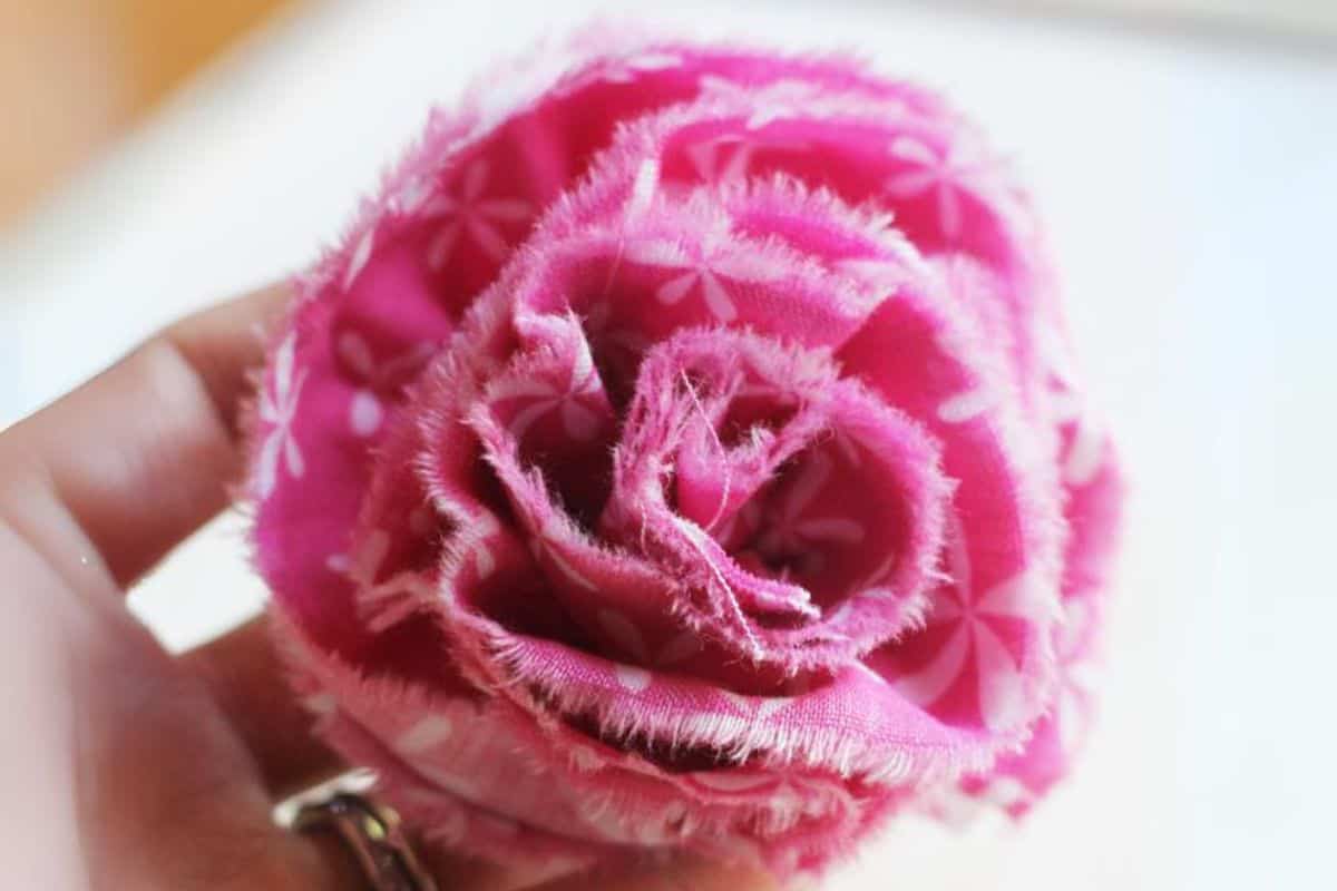Close-up of a handmade ruffled fabric flower with frayed edges, held between fingers, showing layered petals and stitched details.