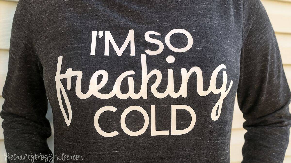 Close-up of a person wearing a long sleeve sweatshirt with a humorous winter phrase printed on the front, photographed outdoors against a house exterior.