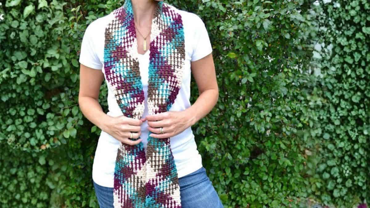 Person wearing a handmade crochet scarf with color pooling, shown draped around the neck and held at the ends while standing outdoors in front of greenery.