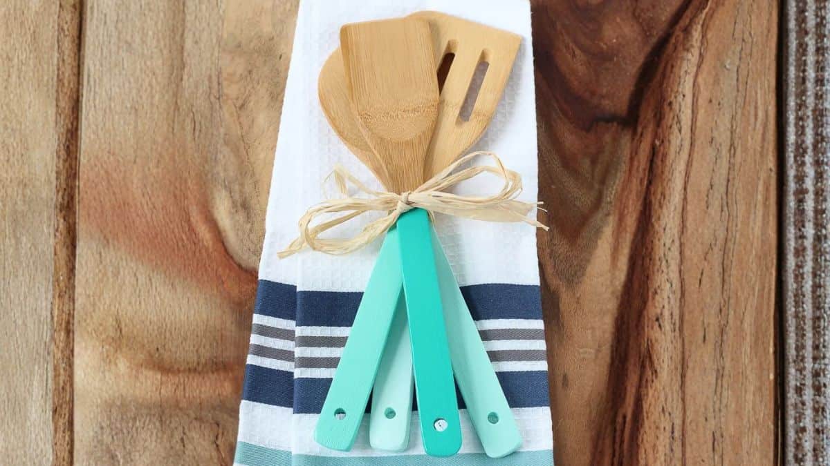 Bamboo kitchen utensils with painted handles tied together and displayed on a folded striped dish towel on a wooden surface.