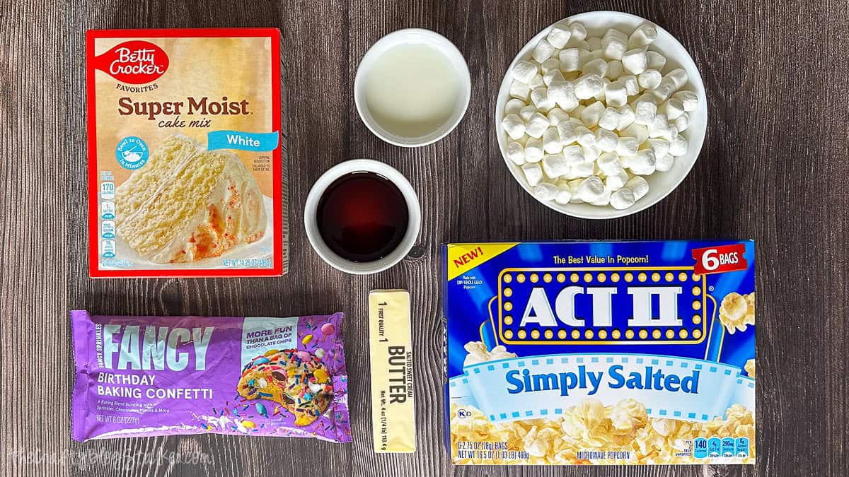 Ingredients for a popcorn cake arranged on a table, including cake mix, microwave popcorn, marshmallows, butter, vanilla, sweetened condensed milk, and confetti sprinkles.