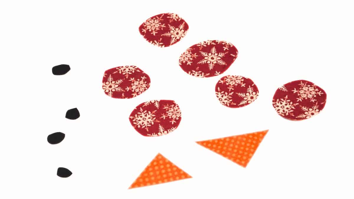 Cut paper pieces for a snowman craft, including small black shapes for eyes and mouth, patterned circles for buttons, and triangle shapes for carrot noses.