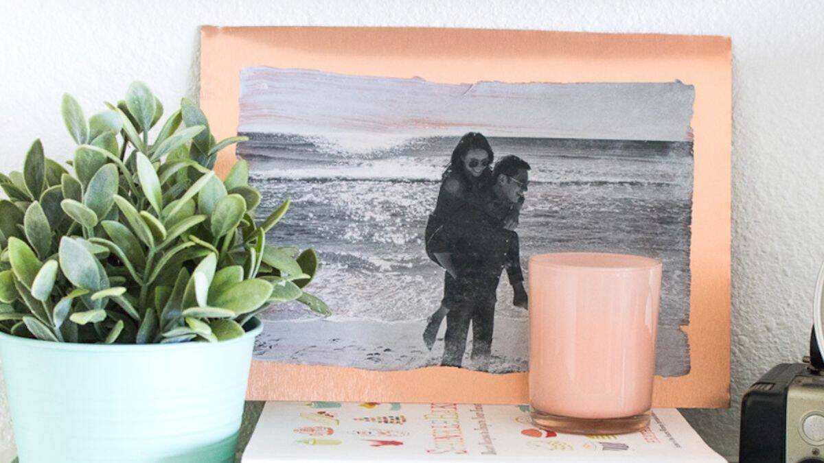 A metal photo transfer of a couple at the beach displayed on a shelf with a potted plant, candle, vintage camera, and stacked books.