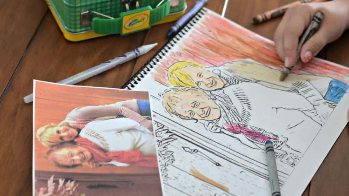 A child colors a custom coloring page created from a printed photo of two smiling people, with crayons and the original photo beside the sketchbook.