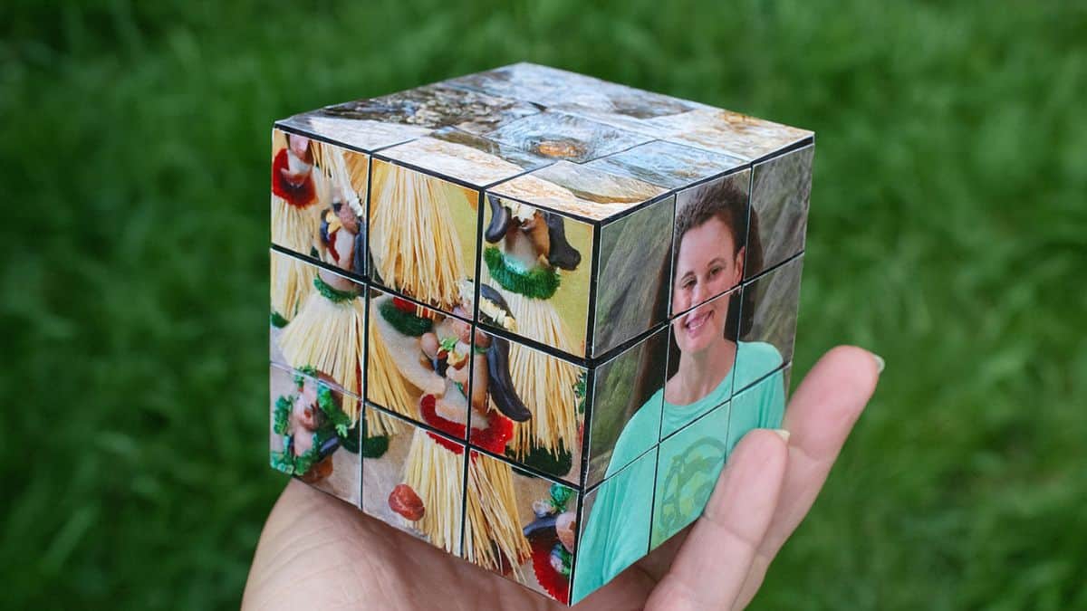 Hand holding a personalized photo cube made from small square tiles, featuring family photos arranged like a Rubik's cube against a grassy background.