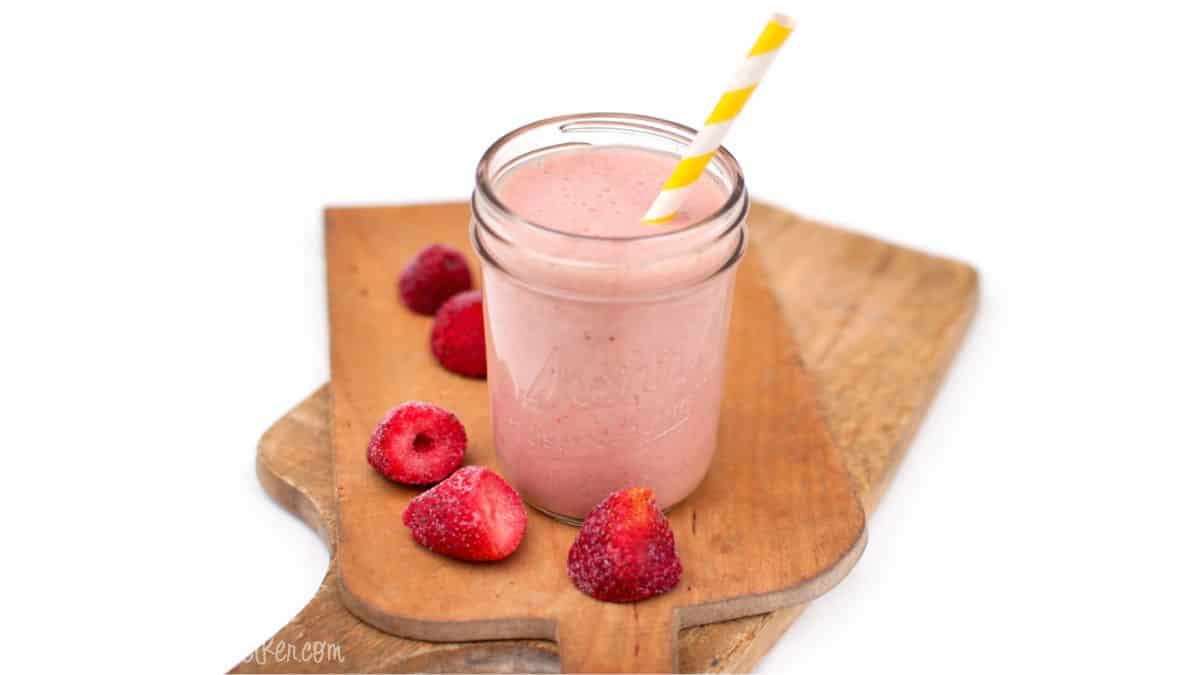 Strawberry smoothie in a mason jar with a paper straw, sitting on a wooden cutting board with whole strawberries beside it.