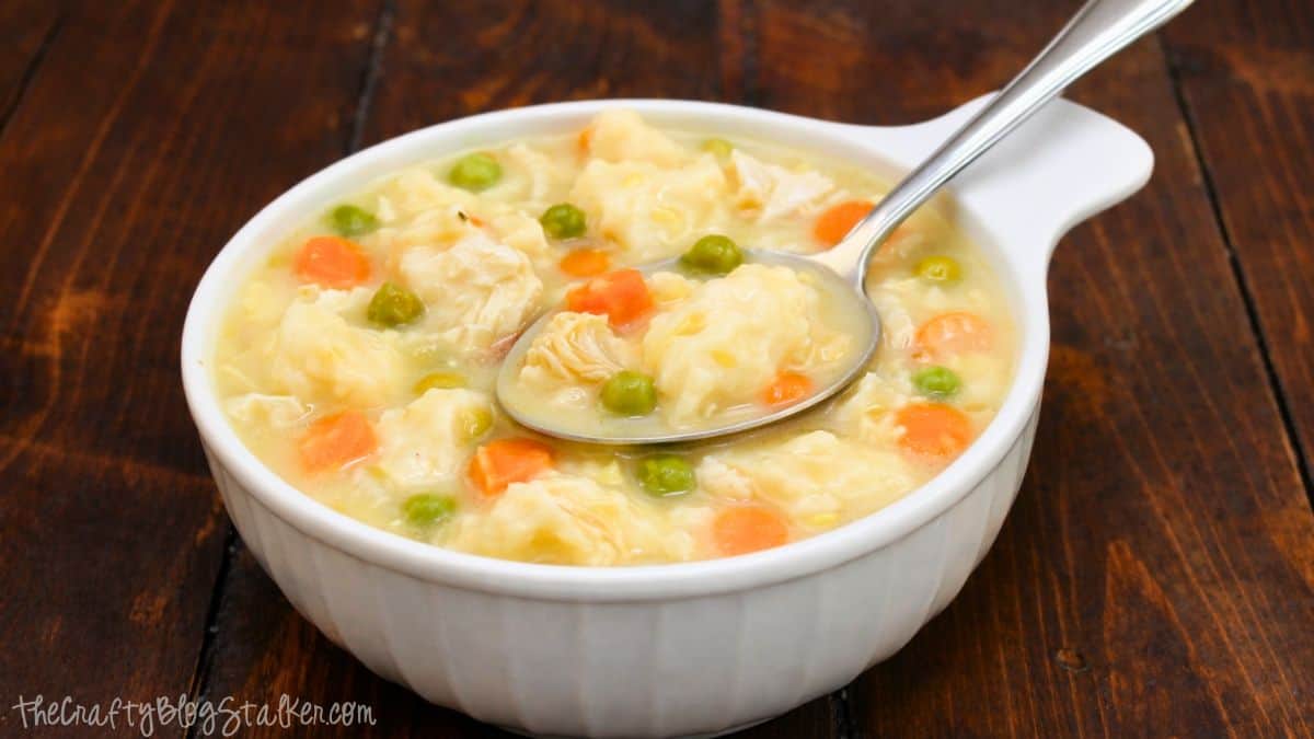 Homemade chicken and dumplings in a white bowl with tender chicken, fluffy dumplings, peas, and carrots in a creamy broth on a wooden table