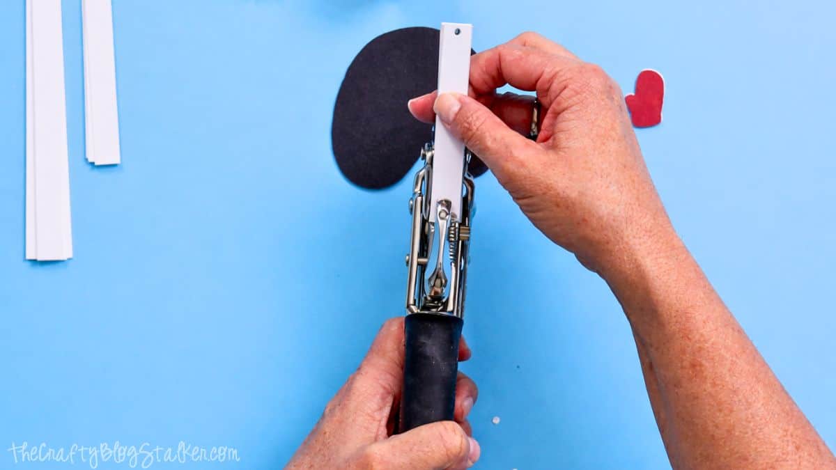 Hands using a Crop-A-Dile tool to punch a hole in a narrow paper strip, with snowman craft pieces and paper strips on a work surface.