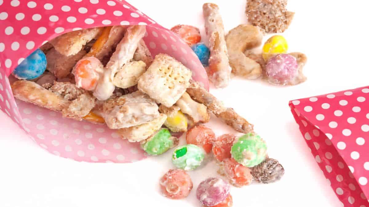 White chocolate party mix with cereal, pretzels, and candy spilling from pink polka dot paper cones onto a white surface.